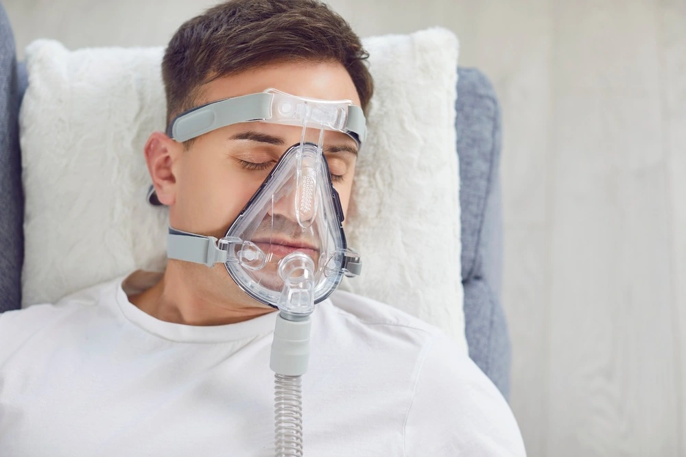 Closeup from above sick man with a medical oxygen mask on face lying on a soft pillow on the couch at home.