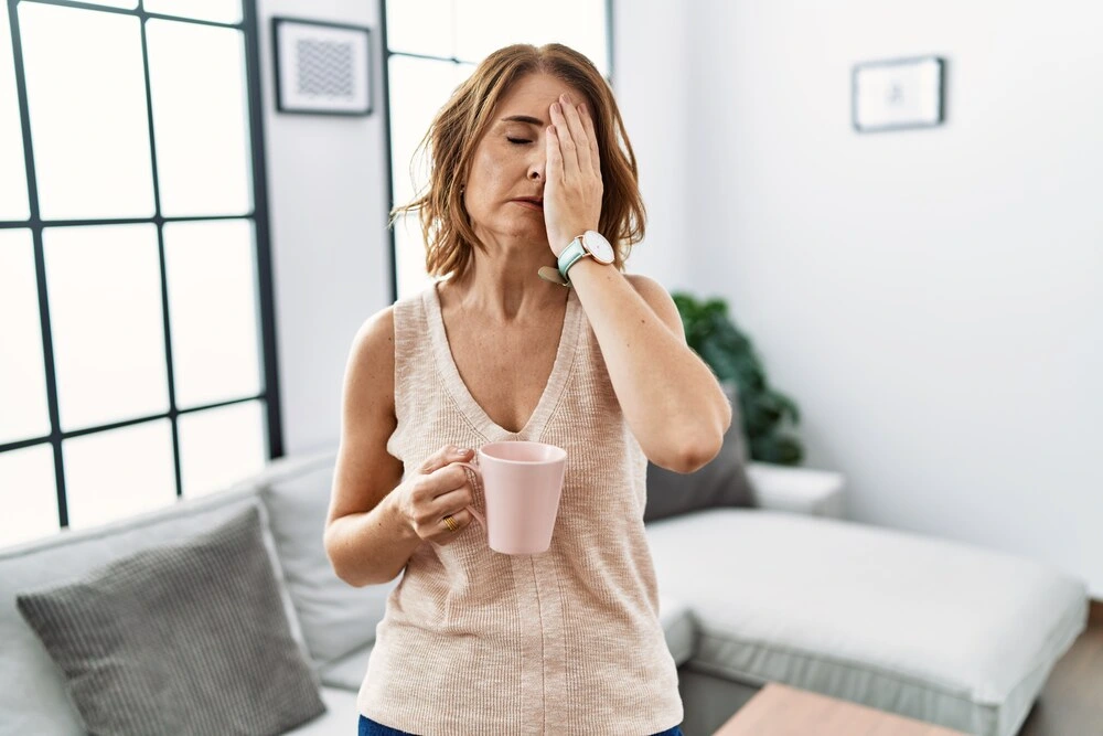 Middle age woman drinking a cup coffee at home yawning tired covering half face, eye and mouth with hand.
